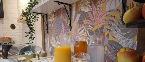 A breakfast bar with various beverages and food items, including a pitcher of orange juice, a glass of milk, and a bottle of iced tea, set against a wall with a floral pattern.