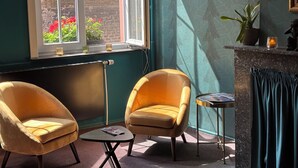 Una habitación con dos sillones, una mesita, un radiador, una ventana con vistas a un muro de ladrillo y flores rojas, y una chimenea con cortina.