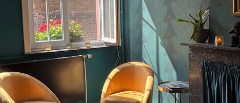 A room with two armchairs, a small table, a radiator, a window with a view of a brick wall and red flowers, and a fireplace with a curtain.