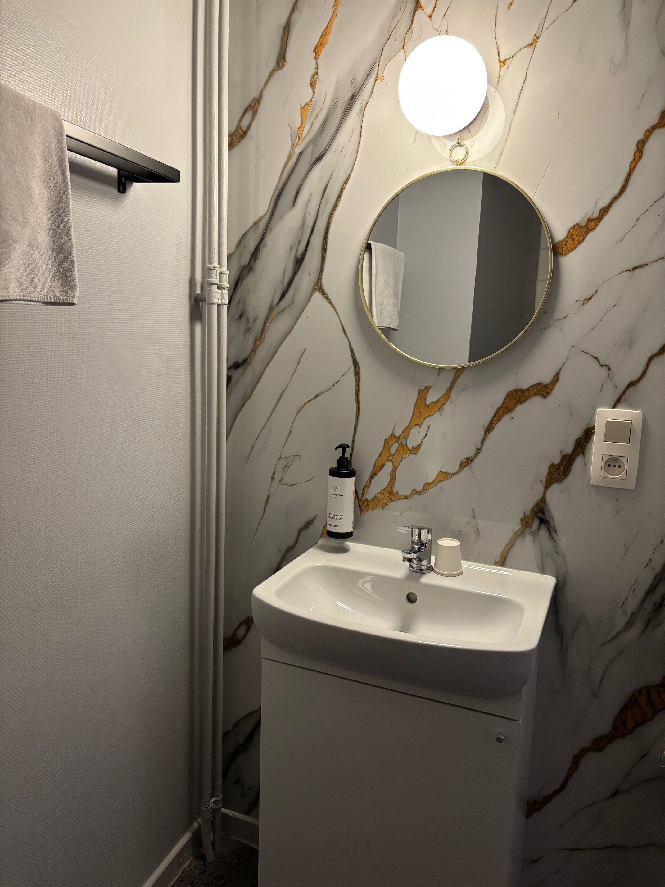 A bathroom with a marble wall, a round mirror, a sink, a towel rack, and a soap dispenser.