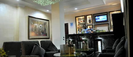 A lobby with dark patterned sofas, a glass coffee table, a chandelier, a bar area with bottles, and a television.