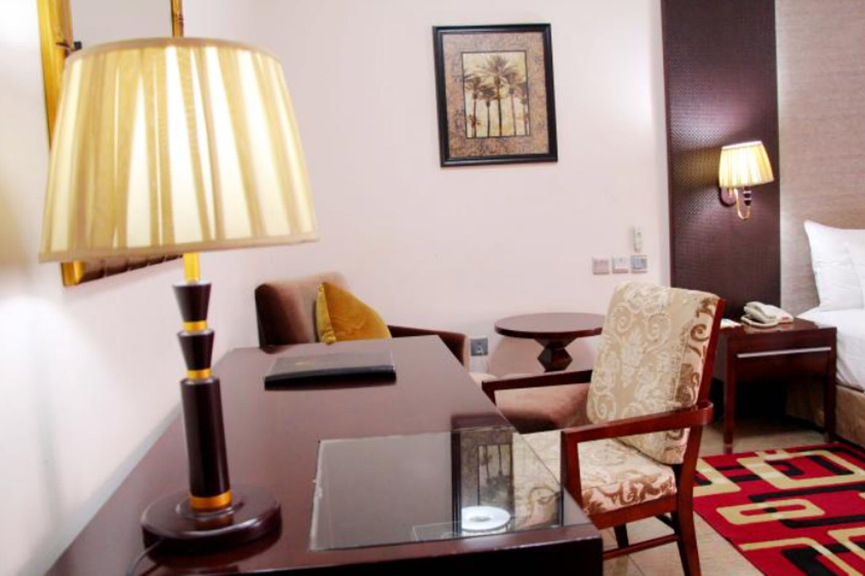A hotel room with a bed, a desk with a lamp, a chair, a small table, and a wall-mounted phone.