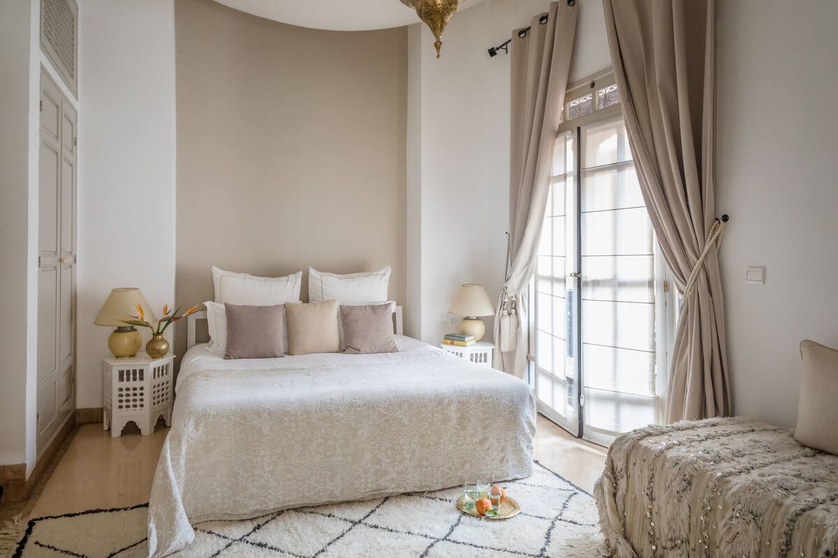 Un dormitorio con una cama grande, una mesita de noche con una lámpara, una pequeña mesa con un florero y una ventana con cortinas.