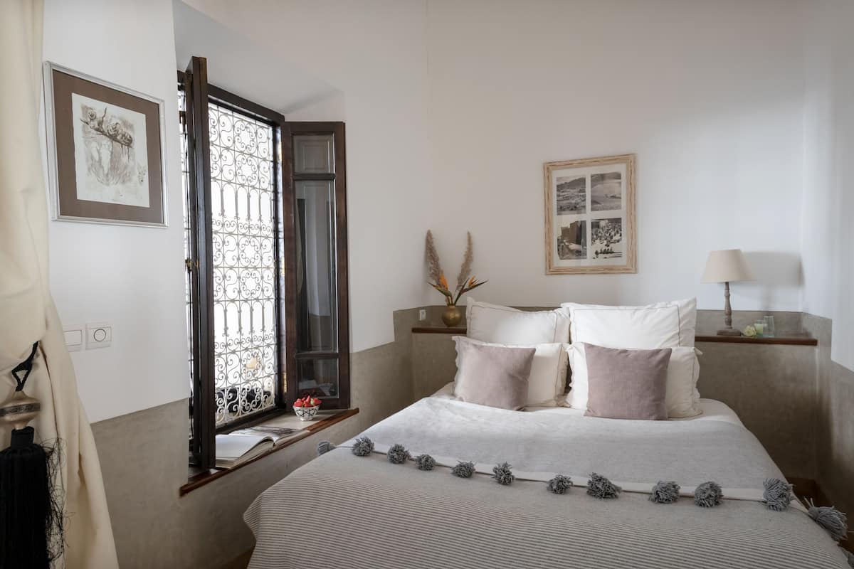 Un dormitorio ordenado con cama, mesitas de noche y ventana con rejas decorativas de hierro.