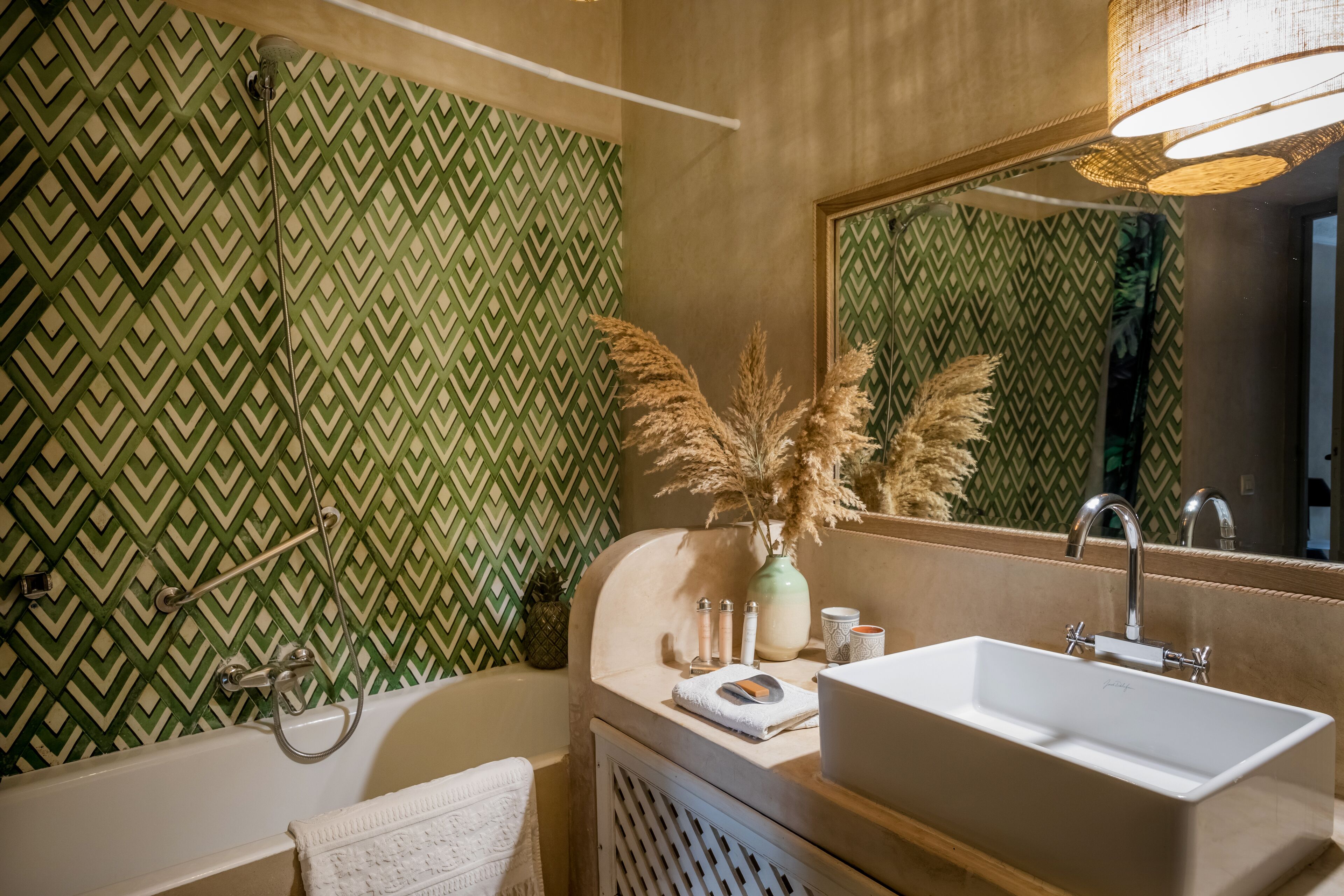Une salle de bain avec un mur à motifs verts, une baignoire blanche, un lavabo avec un miroir et un vase contenant des plantes séchées.