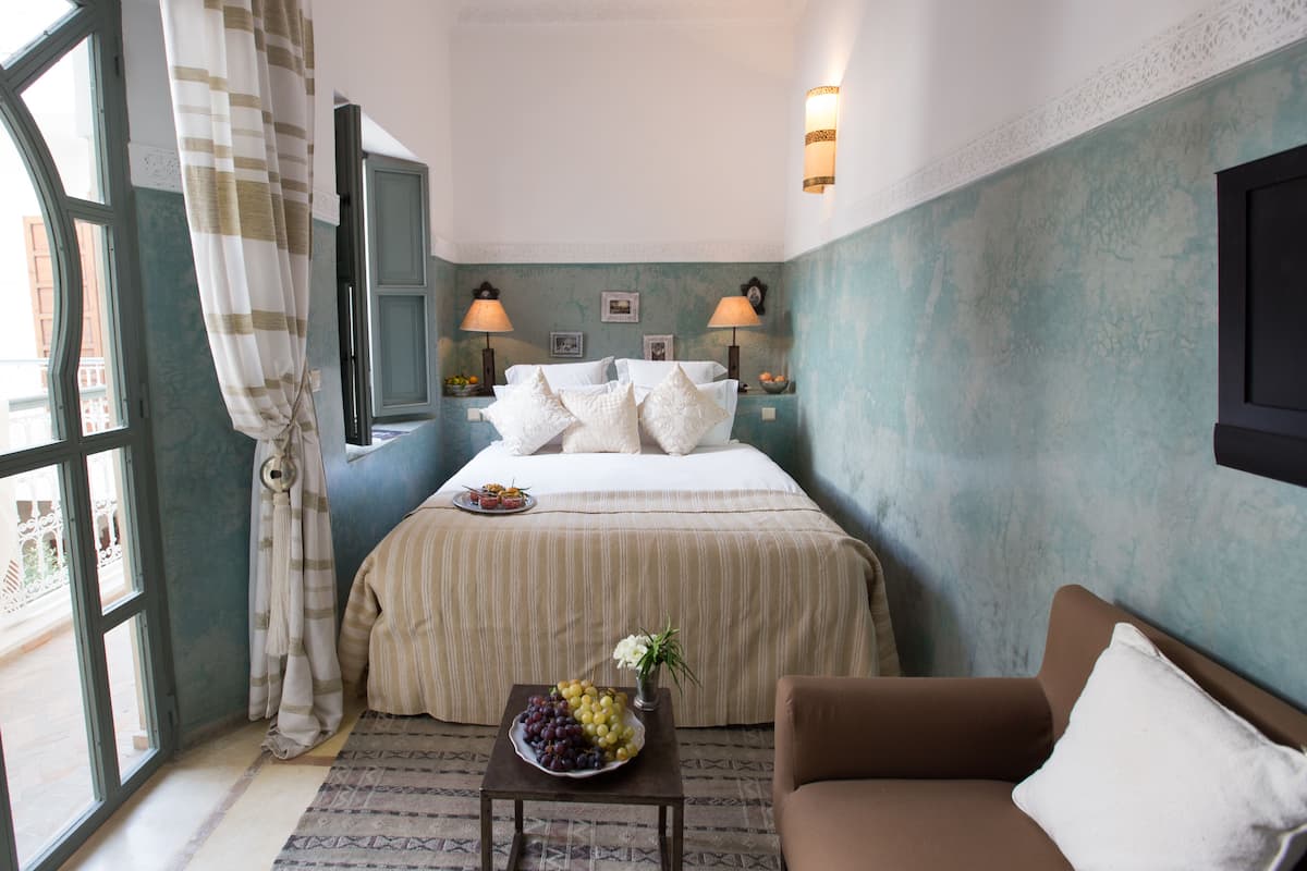 Habitación de hotel con cama, dos lámparas de noche, una mesita con fruta, un sofá marrón y vistas a través de una ventana.