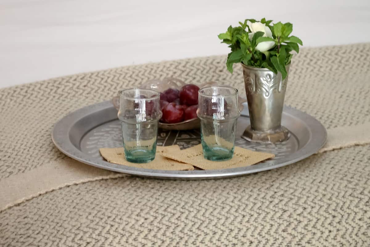 Una cama bien hecha con almohadones estampados, una bandeja con copas y uvas, y una pequeña planta en maceta sobre una alfombra con textura.