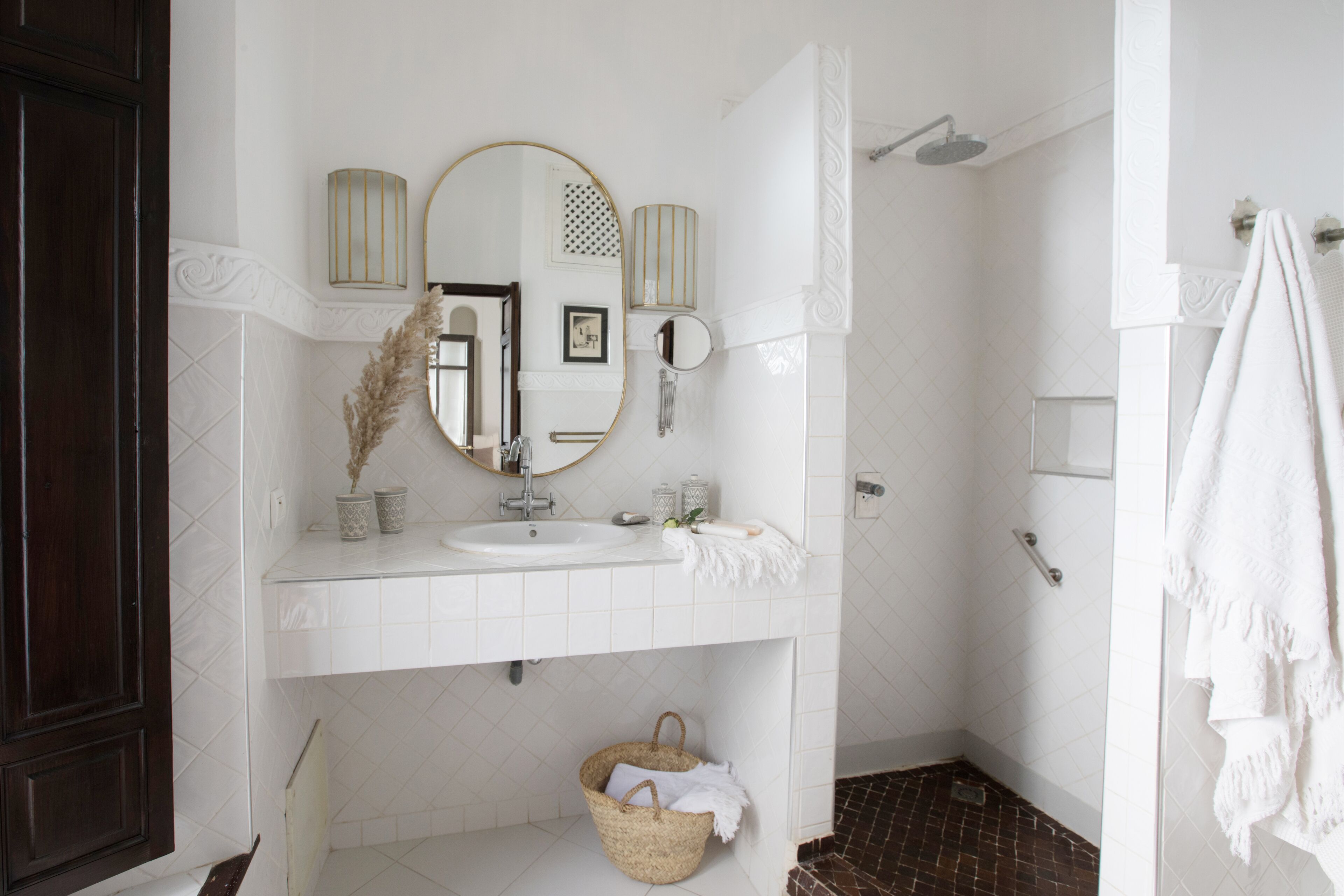 A bathroom with a white sink, mirror, and shower. There is a basket under the sink and a towel hanging on the wall.