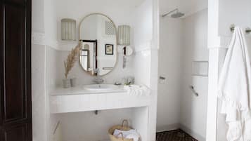 A bathroom with a white sink, mirror, and shower. There is a basket under the sink and a towel hanging on the wall.