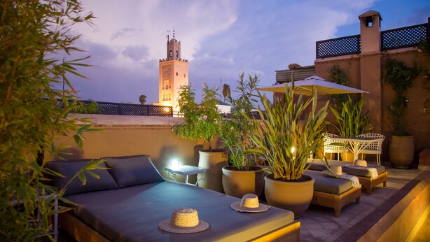 A rooftop terrace with a sofa, chairs, potted plants, and a view of a tower in the background. - Riad Olema et Spa (Marrakech)