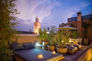 A rooftop terrace with a sofa, chairs, potted plants, and a view of a tower in the background. - Riad Olema et Spa (Marrakech)