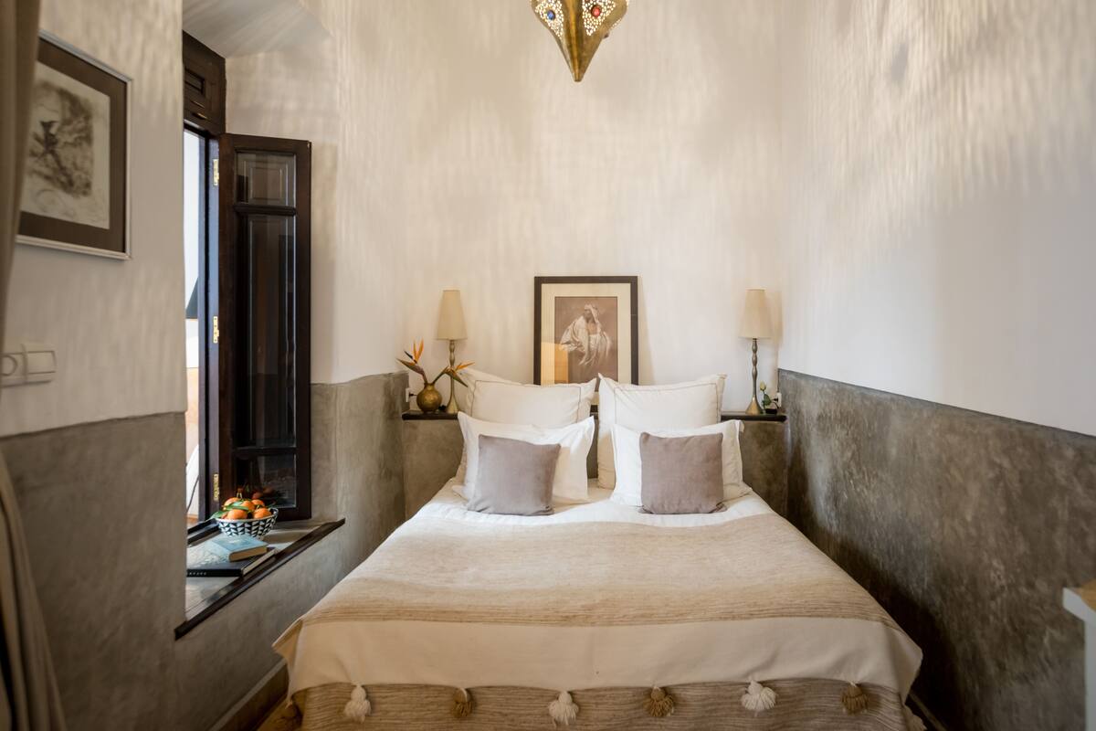 Un dormitorio ordenado con una cama, lámparas de noche, un cuadro enmarcado y una ventana con vistas.