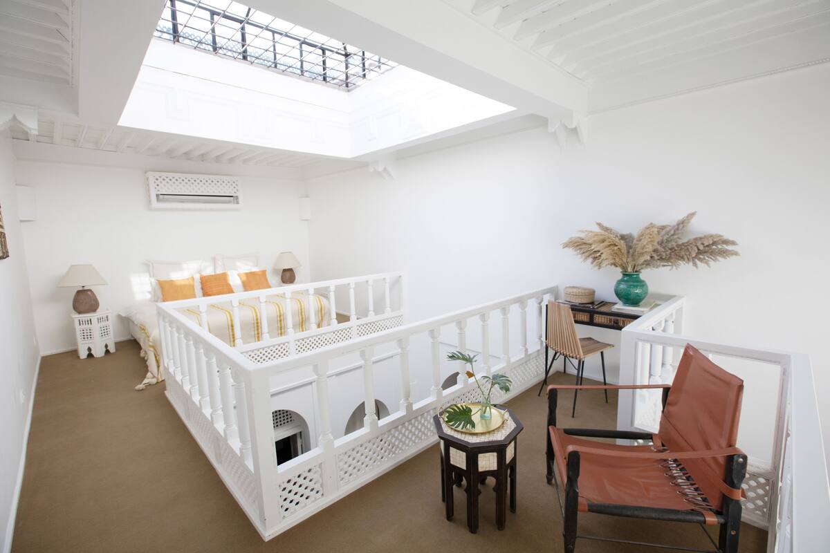 Una habitación luminosa con tragaluz, una cama blanca con almohadas amarillas, una silla de madera y una mesita con una planta.