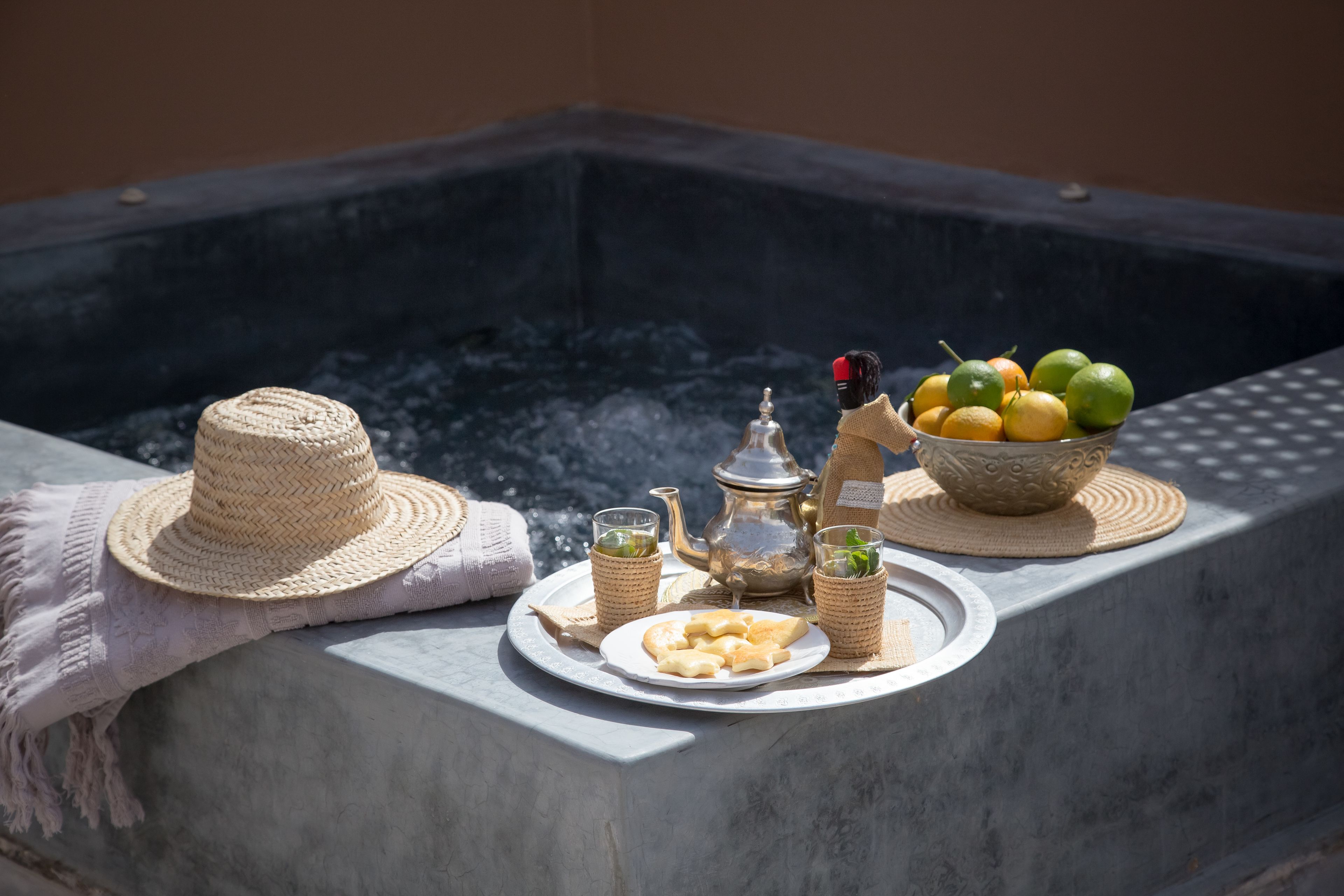 Una vasca idromassaggio con un cappello di vimini, una teiera, bicchieri, un piatto di biscotti e una ciotola di frutta.