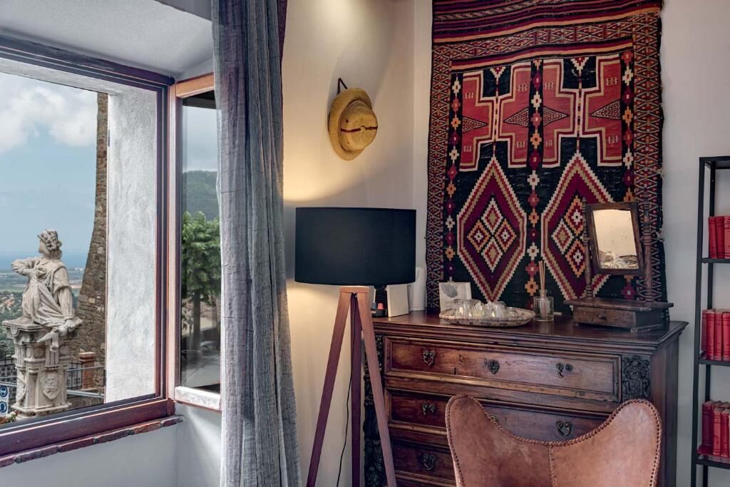A room with a statue by a window, a desk with a lamp, a patterned tapestry, and a bookshelf with books.