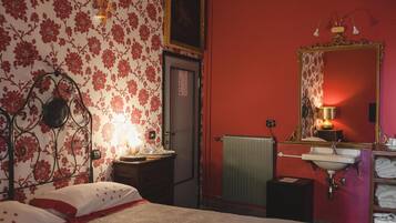 A bedroom with a bed, a bedside table with a lamp, a radiator, a mirror, and a shelf with towels.
