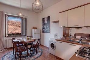 A compact kitchen with a washing machine, a dining area with a table and chairs, and a window with a view of a tiled roof. - Scarlino Ospitalità Diffusa (Scarlino)