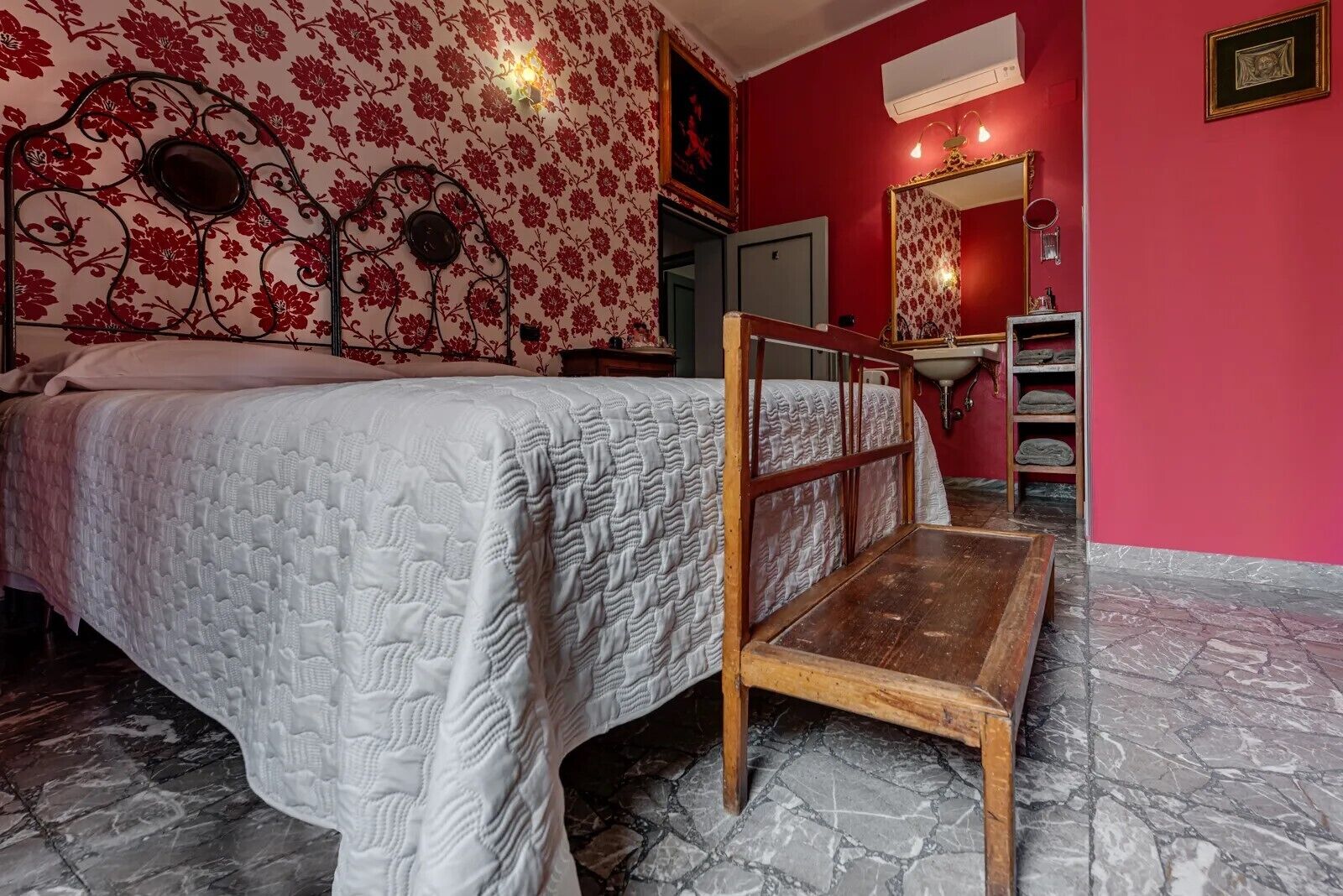 A bedroom with a bed, a wooden bench, a mirror, a sink, and a red wall with floral wallpaper.