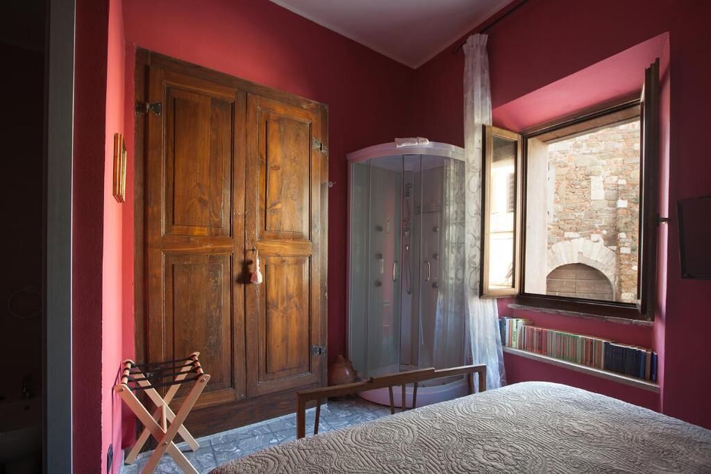 Una stanza con un ampio armadio in legno, una panca, un vetrina e una finestra che si affaccia su un edificio in pietra.