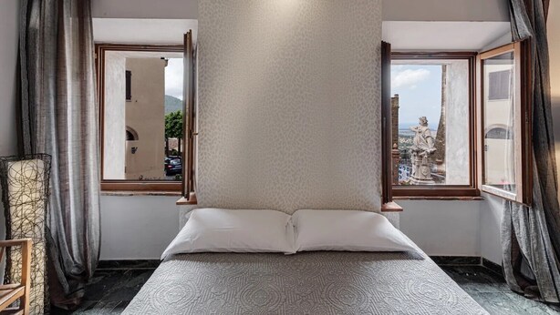 Ein Schlafzimmer mit einem großen Bett, zwei Vorhängen und Fenstern, Blick auf die Stadt und einer Statue vor dem Haus.