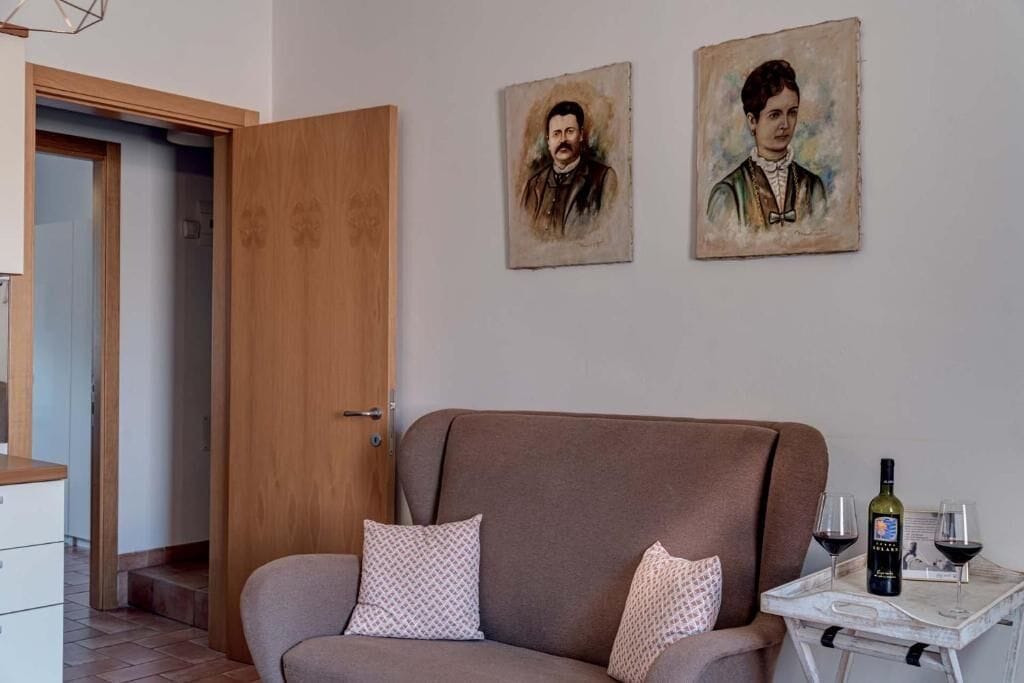 A room with a sofa, two pillows, a painting, and a table with a bottle and glasses.