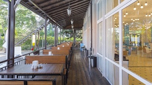 A covered outdoor seating area with wooden tables and benches, large windows, and hanging lights. - MPM Hotel Astoria (Sunny Beach)