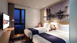 A Travelodge hotel room with two beds, a large window, a painting of a cityscape, and a TV displaying the hotel’s logo.