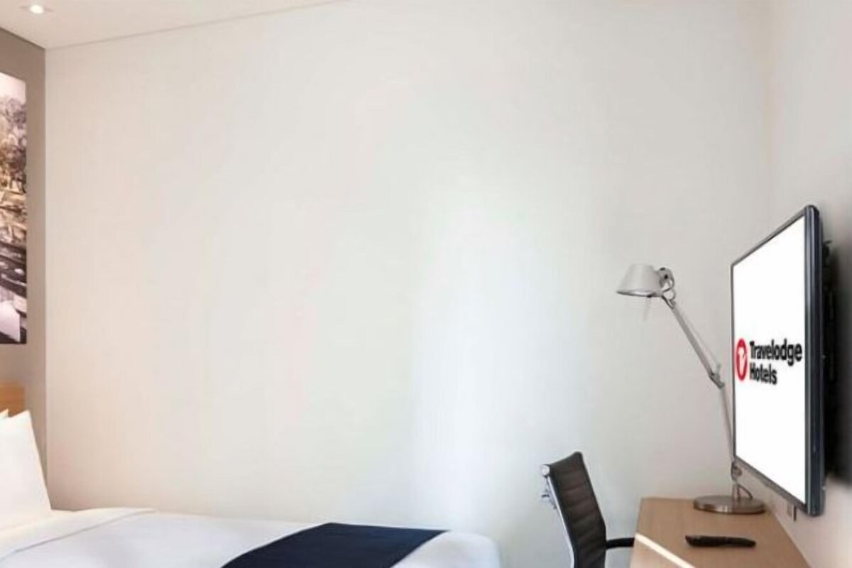 A hotel room with a bed, bedside lamp, desk, and a TV displaying the Travelodge logo.