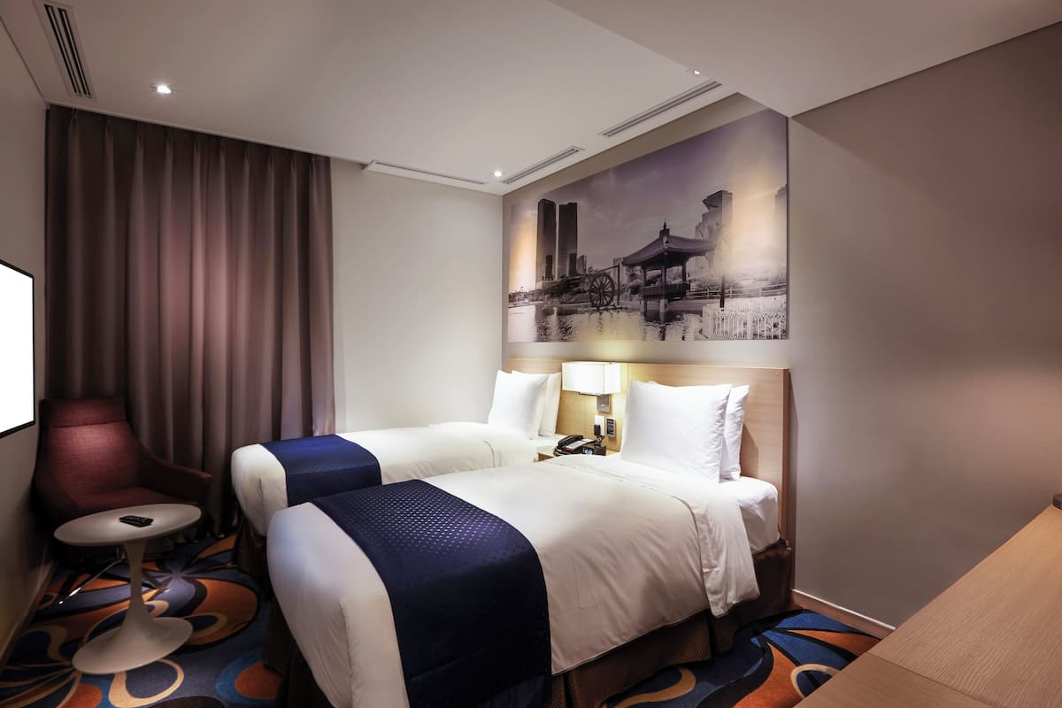 Une chambre d’hôtel avec deux lits, un bureau, une chaise et un grand tableau représentant un paysage urbain accroché au mur.