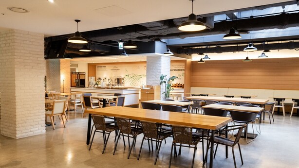 A modern dining area with long wooden tables and wicker chairs, a bar area with a counter, and exposed ductwork on the ceiling.
