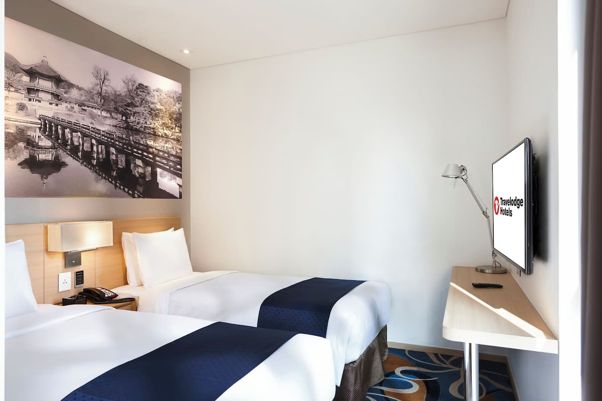 Une chambre d’hôtel avec deux lits, un bureau avec un ordinateur, une télévision et un grand tableau accroché au mur.