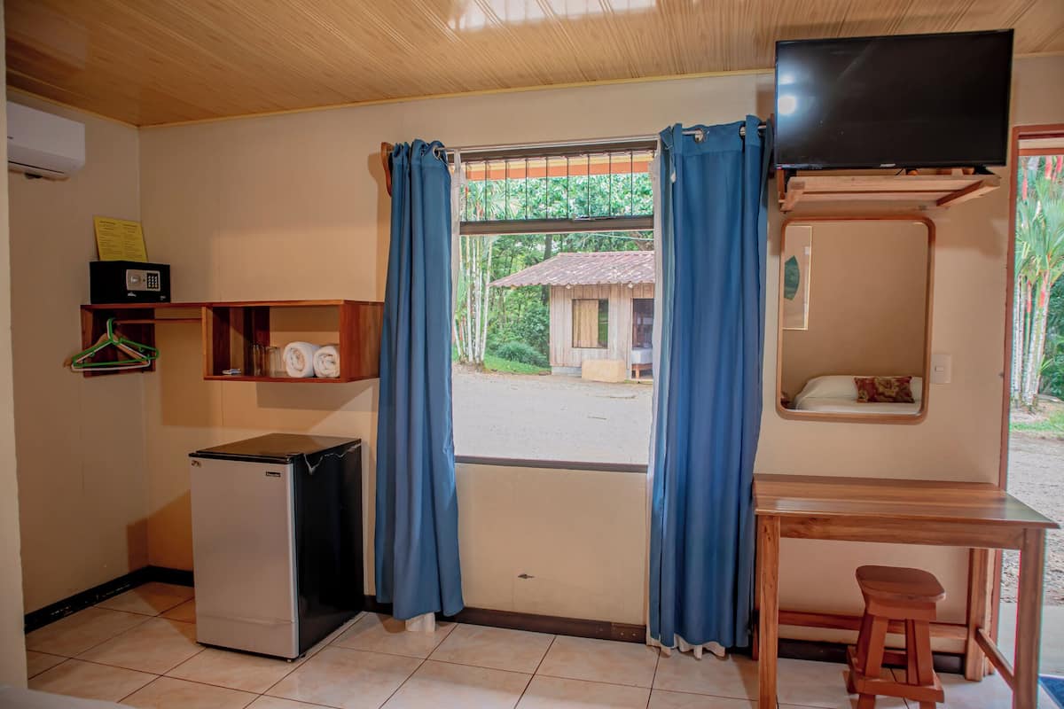 Una habitación pequeña con un refrigerador, una mesa de madera, un televisor montado en la pared y una ventana con cortinas.