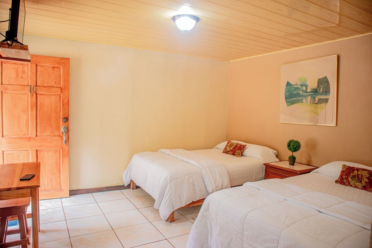 Una habitación con dos camas, un escritorio de madera, una mesita pequeña y una lámpara en el techo.