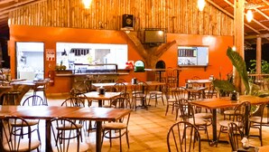 An indoor dining area with wooden walls and ceiling, tables and chairs, a kitchen visible through an open door, and hanging light fixtures. - Hotel del Bosque (Santa Rita)