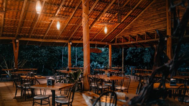 An outdoor dining area with wooden tables and chairs, a thatched roof, and hanging lights. - Hotel del Bosque (Santa Rita)
