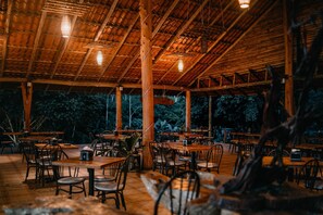 An outdoor dining area with wooden tables and chairs, a thatched roof, and hanging lights. - Hotel del Bosque (Santa Rita)