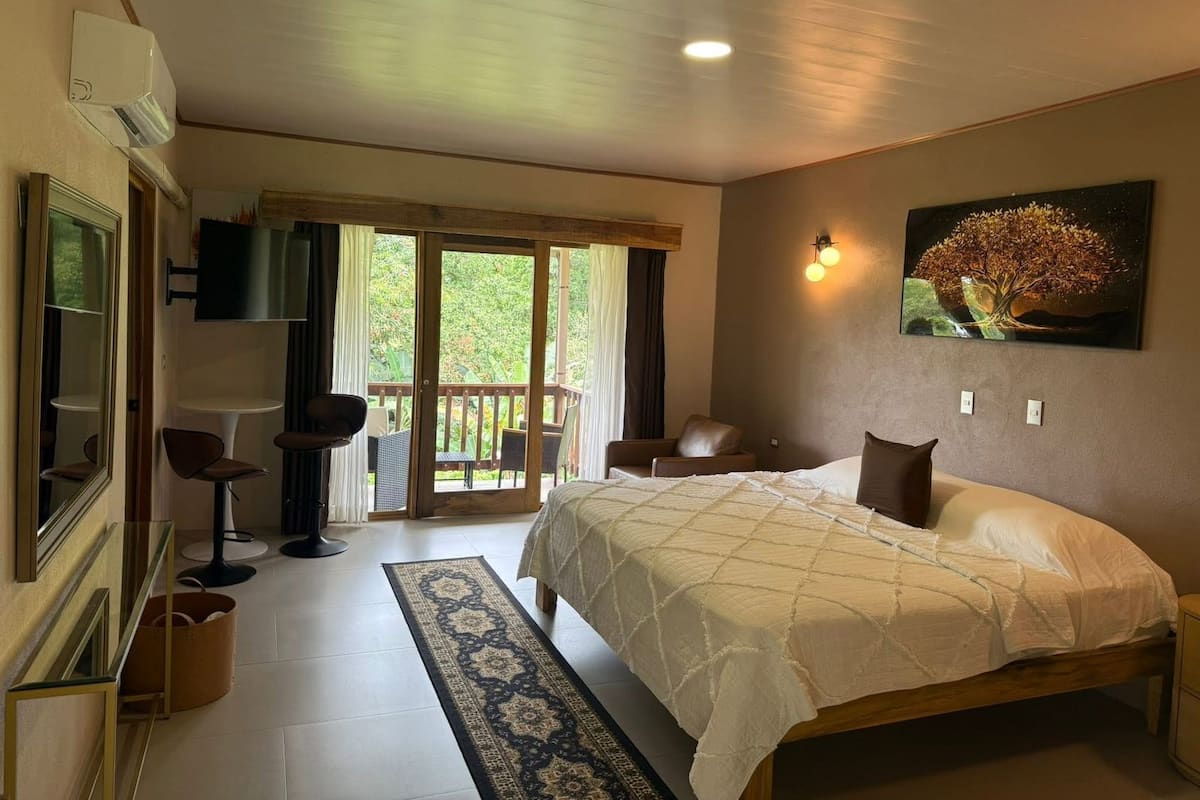 Un dormitorio con cama, televisor, silla, mesa y balcón.