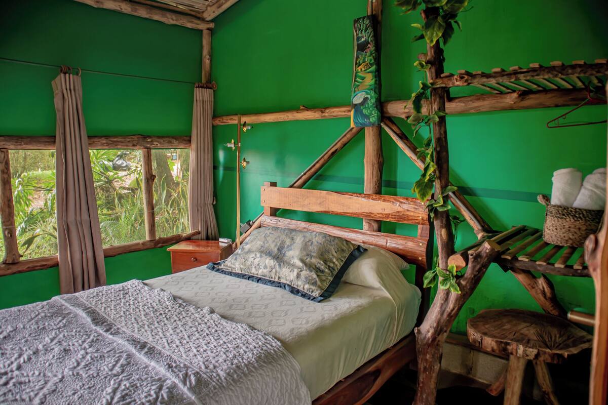 Una habitación rústica con una cama de madera, paredes verdes y una ventana con vistas a la vegetación.