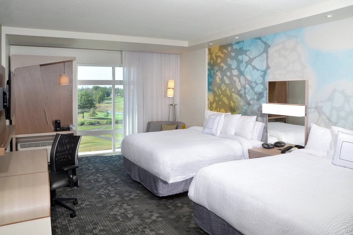 Ein Hotelzimmer mit zwei Betten, einem Schreibtisch, einem Stuhl, einer Lampe und Blick auf einen Golfplatz.