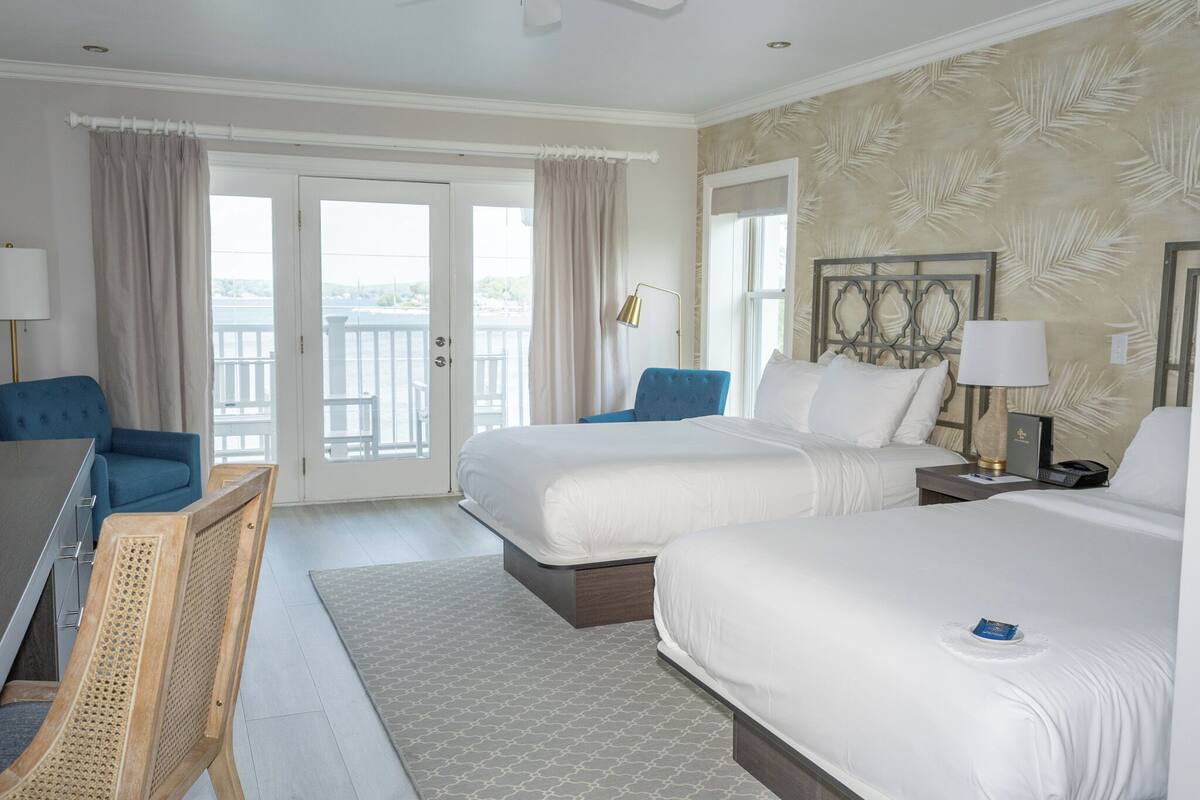 A hotel room with two beds, a desk, a chair, a lamp, and a view of the outdoors.