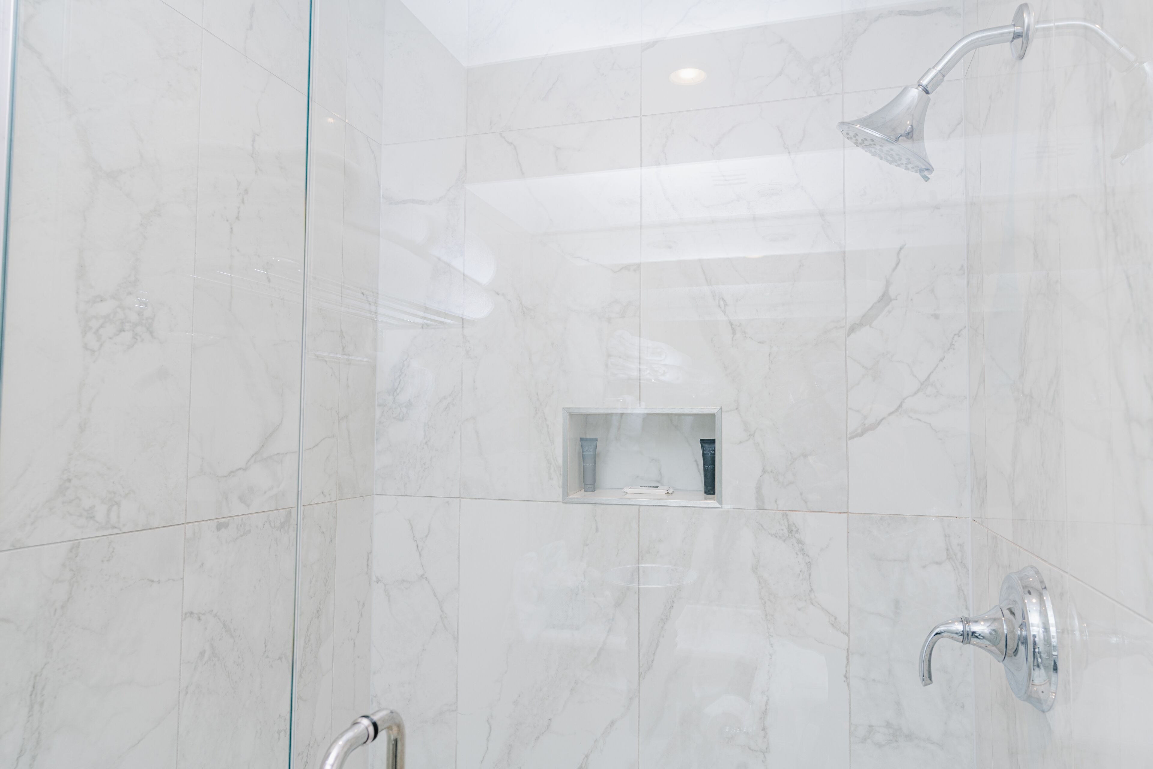 Un baño moderno con cabina de ducha de vidrio, paredes de mármol y un estante empotrado con artículos de aseo.