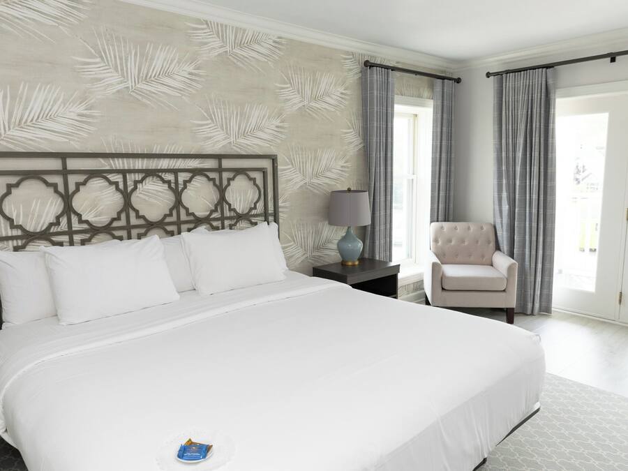 Uma cama grande com cabeceira decorativa, uma mesinha de cabeceira com abajur, uma cadeira e vista para o exterior por uma porta de correr.