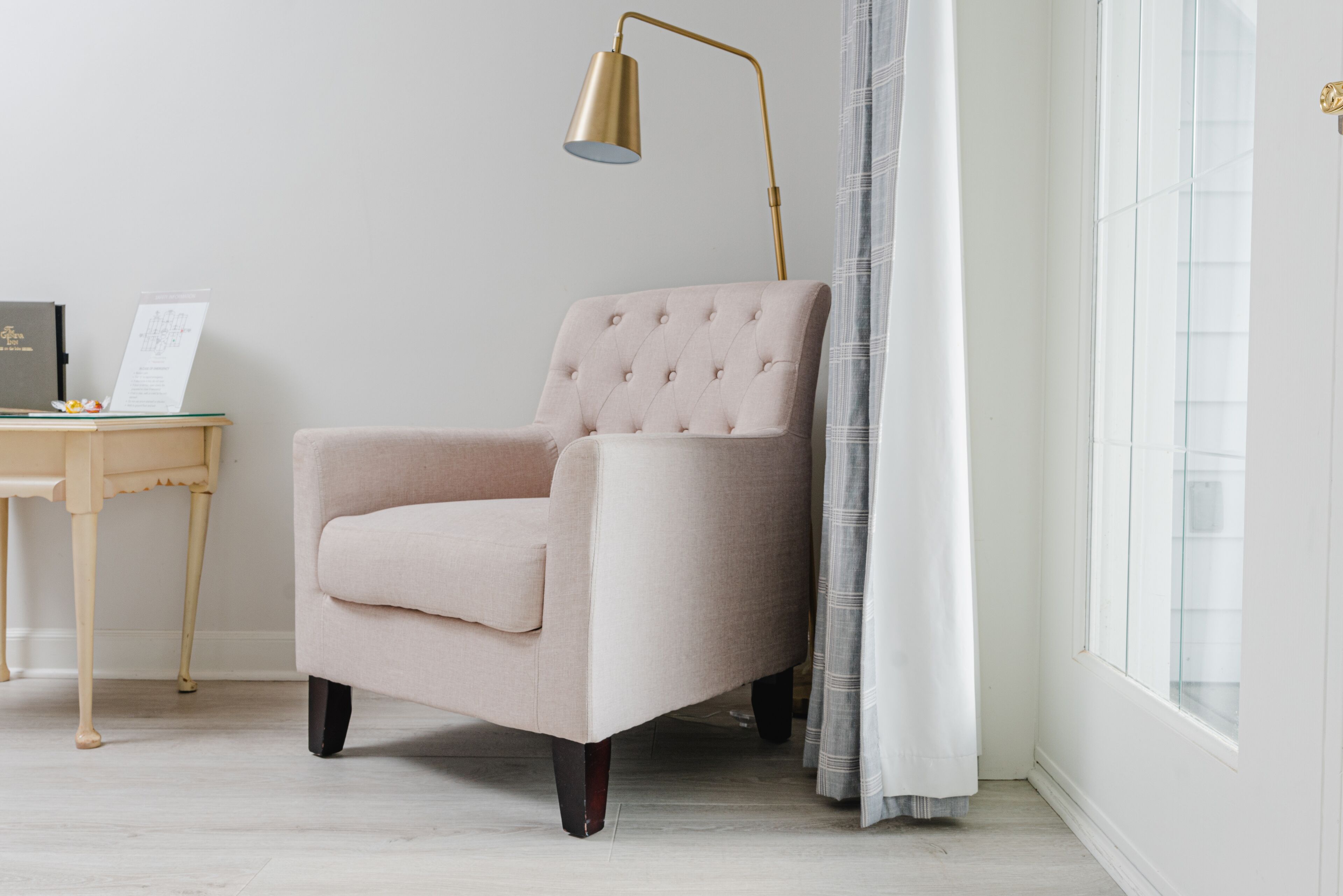 A room with a light pink armchair, a small wooden table with a laptop and papers, a gold floor lamp, and a window with white curtains.