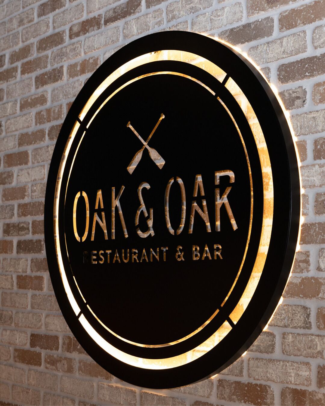 A circular sign with "OAK & OAR" and "RESTAURANT & BAR" text, mounted on a brick wall.