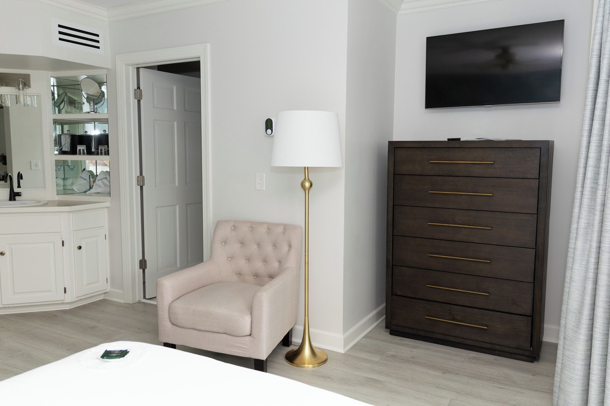 Un baño moderno con un tocador blanco, un sillón beige, una lámpara de pie y un mueble de madera oscura.