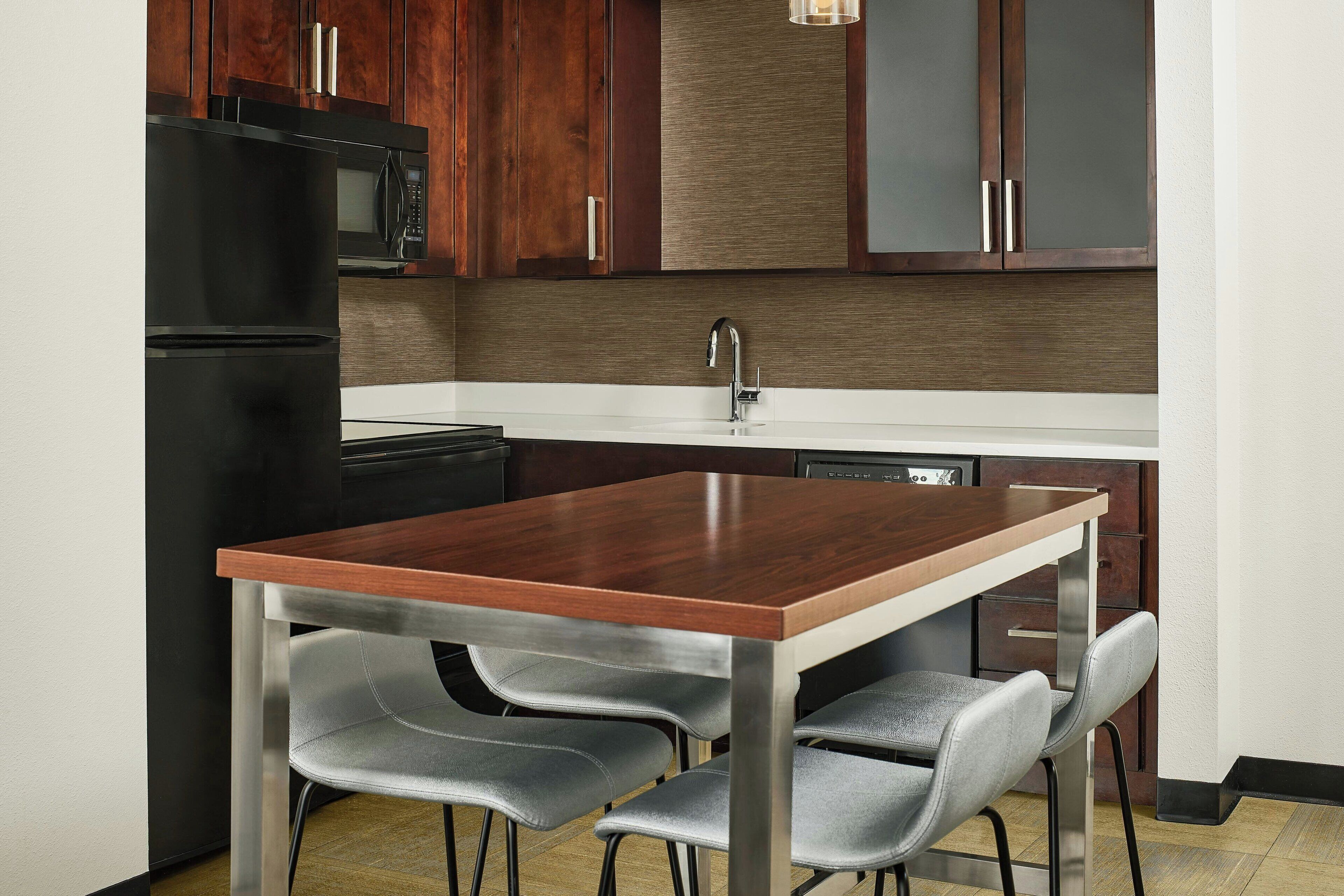 Una cocina compacta con un refrigerador negro, un microondas, un lavamanos de acero inoxidable y una mesa de madera con cuatro sillas.