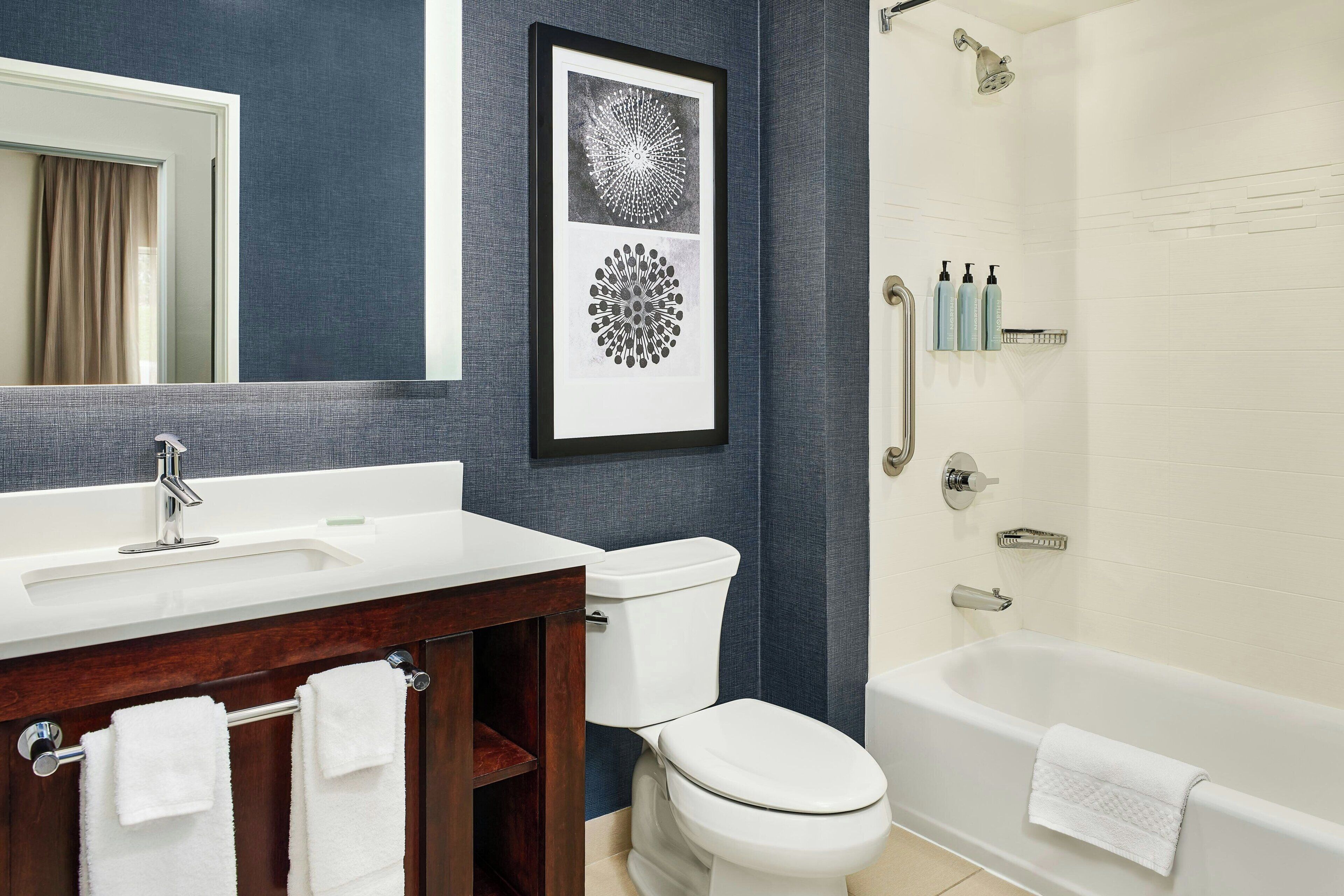 Un baño con lavamanos blanco, espejo, inodoro y bañera, con una obra de arte enmarcada sobre el espejo, y una toalla.