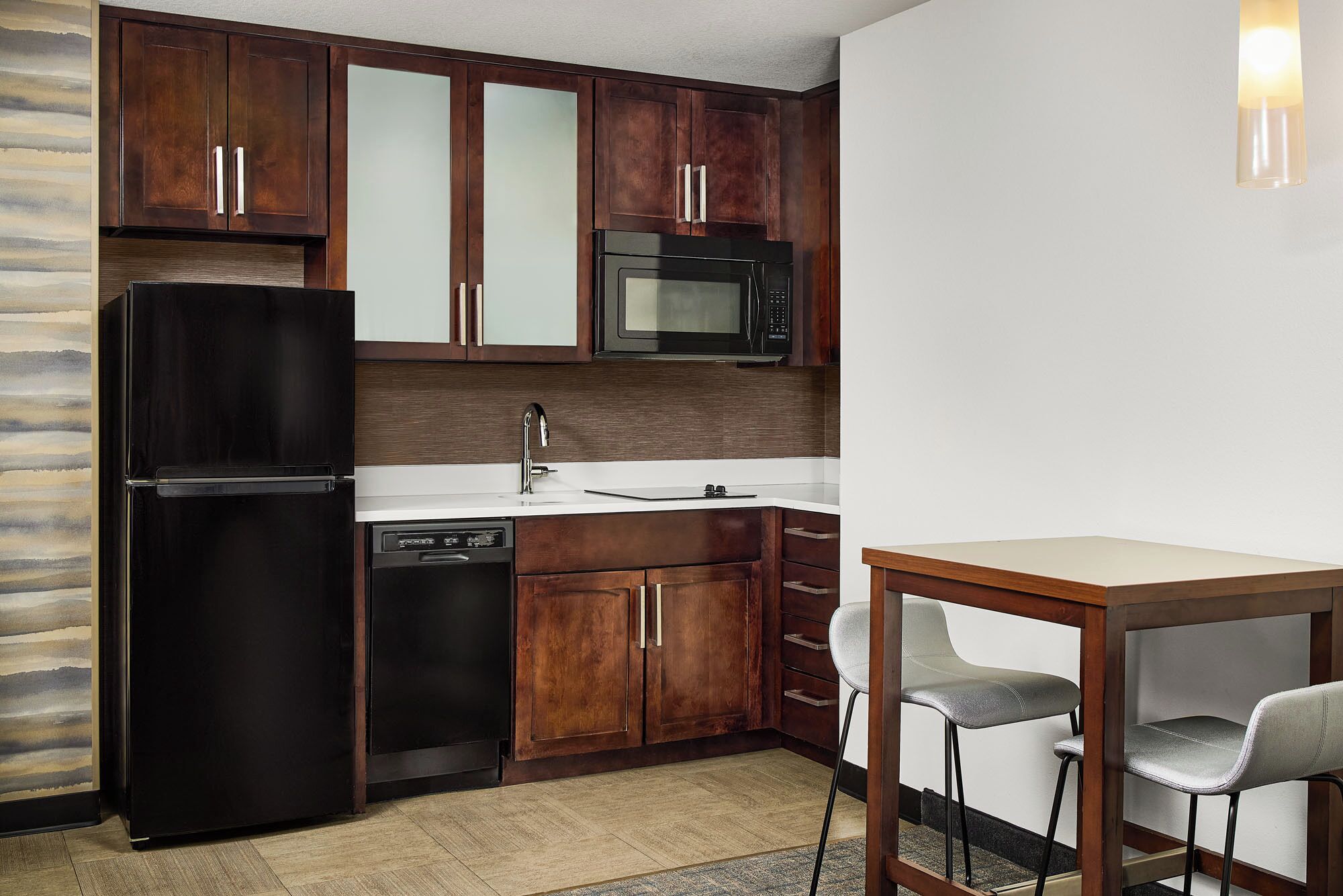 Une cuisine compacte avec des meubles de rangement en bois foncé, un réfrigérateur noir, un four à micro-ondes et un lave-vaisselle. On y trouve une petite table en bois avec deux chaises.