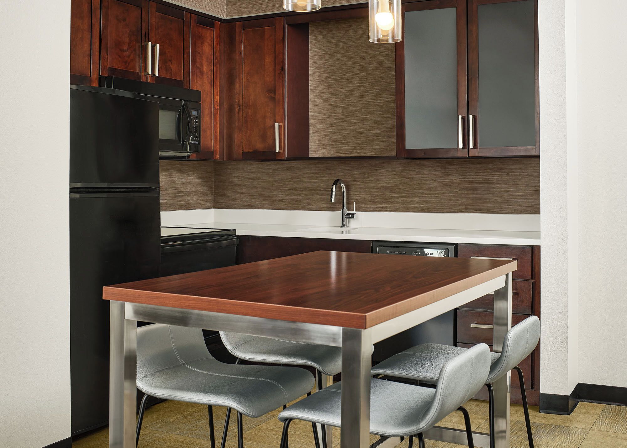Une cuisine compacte avec des meubles de couleur bois foncé, un réfrigérateur noir, un four à micro-ondes en acier inoxydable et un plan de travail blanc. On y trouve une table à manger en bois, accompagnée de quatre chaises grises à revêtement.