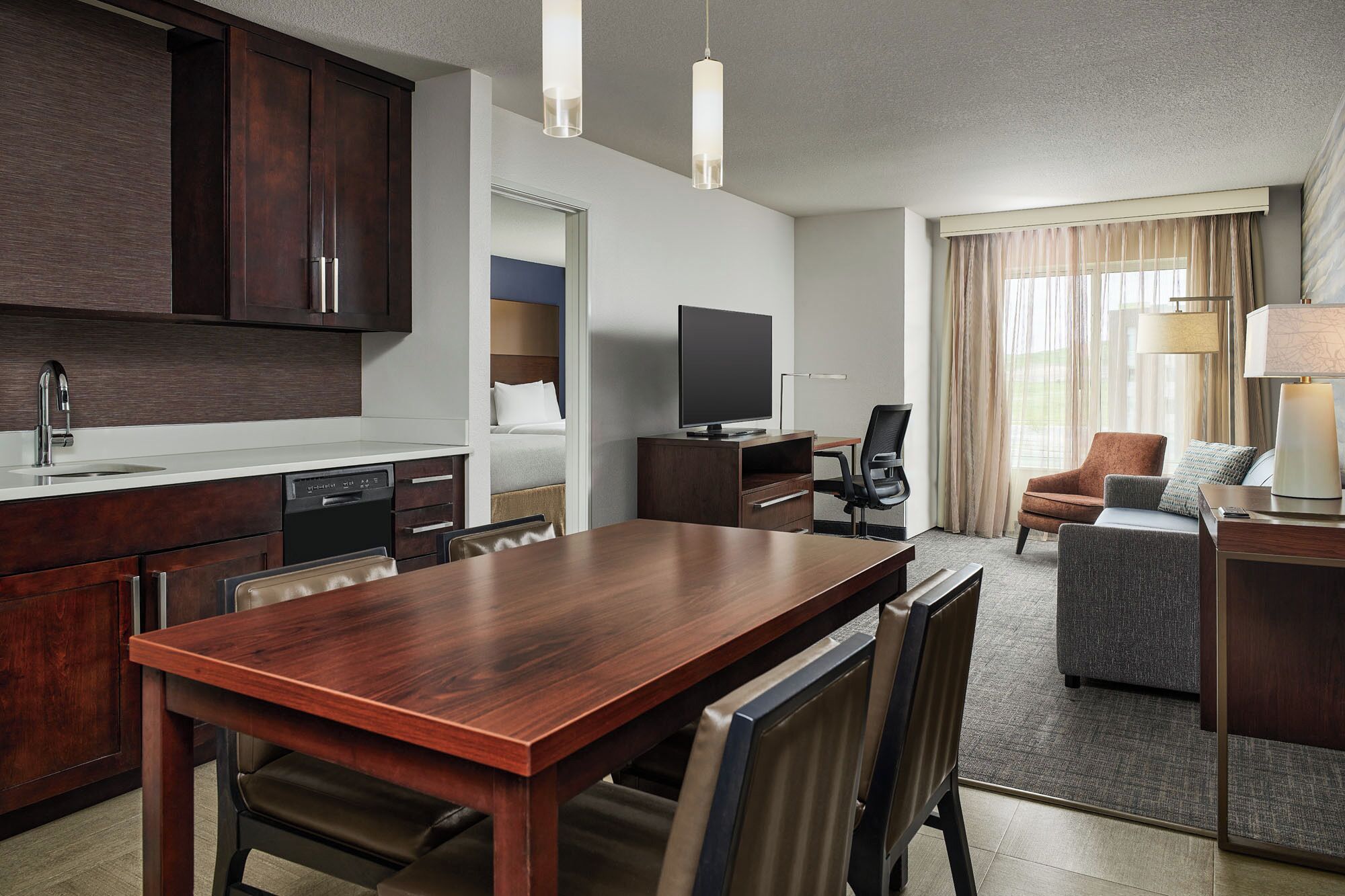 Una habitación de hotel moderna con una mesa de comedor de madera, un televisor de pantalla plana, un sofá y una lámpara.